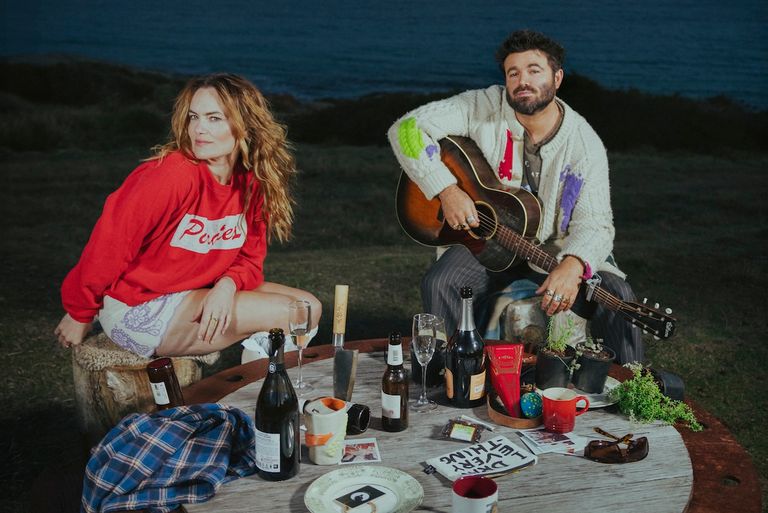 Australian music duo Angus and Julia Stone outdoors at dusk with an acoustic guitar and drinks on a wooden table near the ocean.